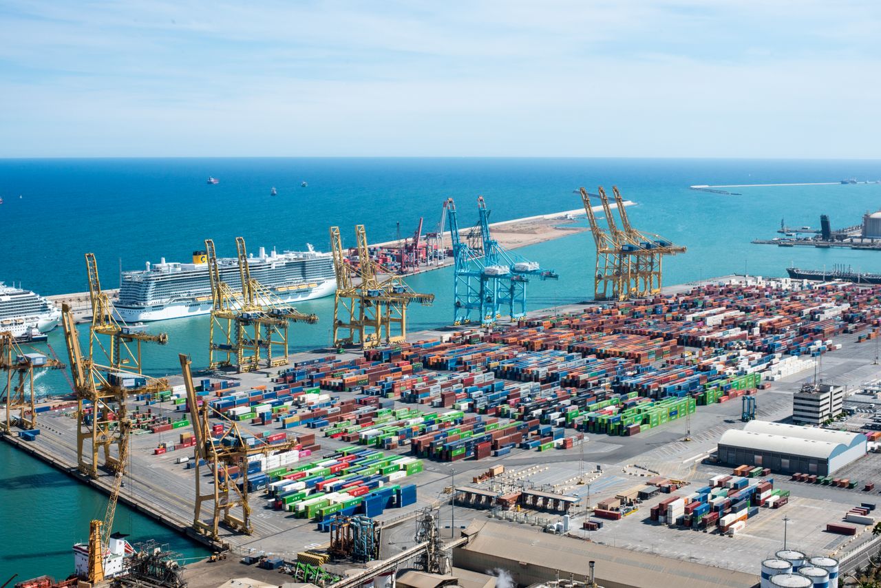 Beranda A vibrant aerial shot of Barcelona port featuring cranes, containers, and cruise ships on a sunny day.